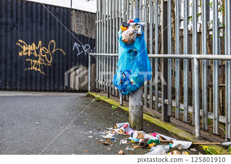 Blue garbage bag hanging on fence with litter scattered on ground in urban area 125801910