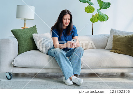Serious young woman sitting at home on the couch using smartphone Serious young woman sitting at home on the couch using smartphone 125802306