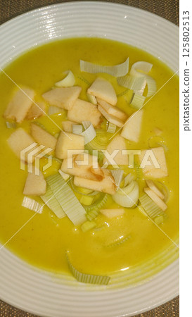 Creamy yellow soup garnished with chopped apples and leeks in white bowl 125802513