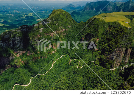 Winding mountain road through lush green forest with dramatic cliffs and panoramic view from above 125802670