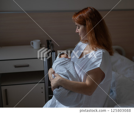 Caucasian woman holding newborn son while sitting on couch in maternity hospital ward.  125803088