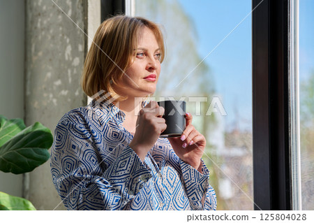 Portrait of beautiful middle-aged woman with cup in her hands looking out window Portrait of beautiful middle-aged woman with cup in her hands looking out window 125804028