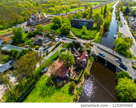 Aerial view of a railway crossing with cars and cyclists, a small station nearby, surrounded by lush green forest under a clear blue sky. Peaceful rural transportation scene. 125804060