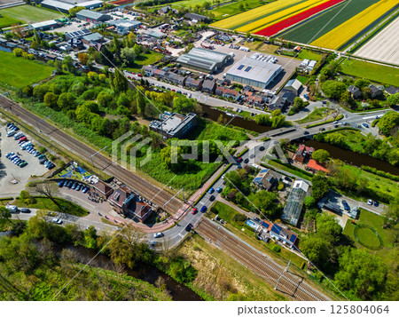Aerial view of a railway crossing with cars and cyclists, a small station nearby, surrounded by lush green forest under a clear blue sky. Peaceful rural transportation scene. Aerial view of a railway crossing with cars and cyclists, a small station nearby, surrounded by lush green forest under a clear blue sky. Peaceful rural transportation scene. 125804064