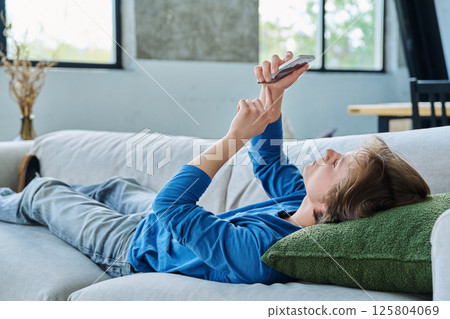Calm serious handsome teenager guy using smartphone lying on the couch at home Calm serious handsome teenager guy using smartphone lying on the couch at home 125804069