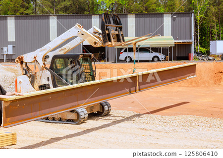 Construction worker operates forklift skid steer loader, moving large steel beam at building site 125806410
