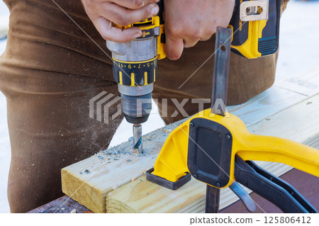 Skilled worker uses power drill to create holes in wooden beam at construction site during works day 125806412
