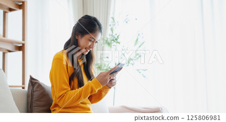 Smiling pretty young Asian woman sitting on cozy couch, using funny mobile apps in living room Smiling pretty young Asian woman sitting on cozy couch, using funny mobile apps in living room 125806981