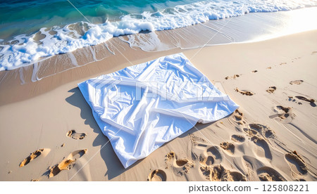 wrinkled white blanket is spread out on sandy beach, with ocean surf gently lapping nearby. Footprints in sand add sense of tranquility and relaxation to scene 125808221