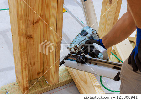 Construction worker skillfully operates nail gun, securing wooden beams to frame structure at busy works site. Construction worker skillfully operates nail gun, securing wooden beams to frame structure at busy works site. 125808941