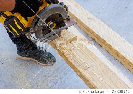 Carpenter works with circular saw to cut wooden plank at construction site during works day 125808942