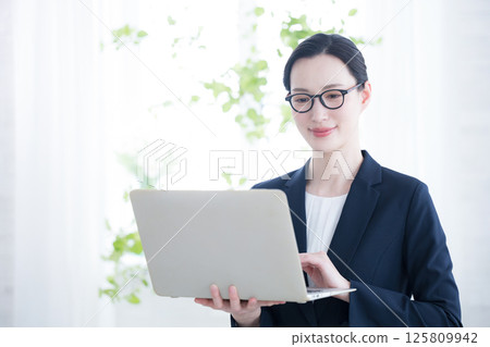 A woman wearing glasses holding a laptop in a beautiful backlit office, organizing tasks, checking schedules, etc. 125809942