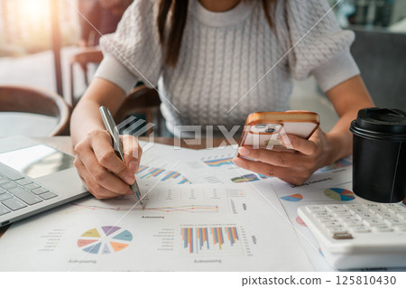 Professional woman reviewing financial charts and using a smartphone, showcasing multitasking in a business environment. Professional woman reviewing financial charts and using a smartphone, showcasing multitasking in a business environment. 125810430