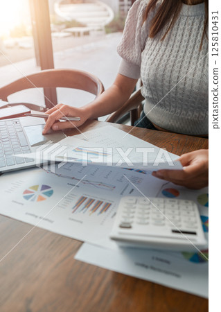 Professional woman reviewing financial documents with charts and calculator on a wooden desk in a bright office. 125810431