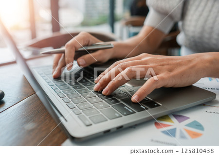 Close-up of hands typing on a laptop in a bright office setting with financial documents. Close-up of hands typing on a laptop in a bright office setting with financial documents. 125810438