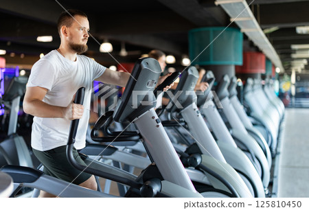 Young man stands on simulator, performs fitness task using an elliptical simulator 125810450
