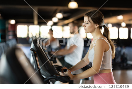 Girl performs exercises with stationary bicycle in gym Girl performs exercises with stationary bicycle in gym 125810489