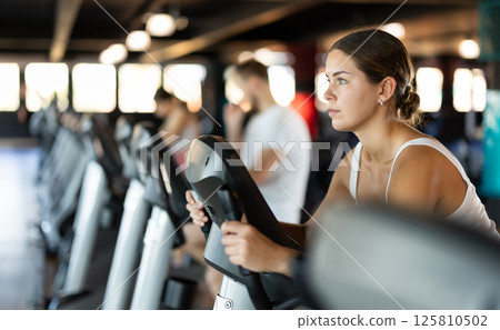 Girl performs exercises with stationary bicycle in gym Girl performs exercises with stationary bicycle in gym 125810502