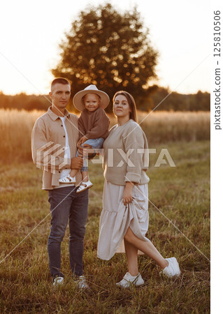 A family of three, a man and a woman with a child in their arms, stand in a field at sunset A family of three, a man and a woman with a child in their arms, stand in a field at sunset 125810506