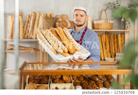 Male seller stands at bakery counter with basket filled by fresh baguettes. Male seller stands at bakery counter with basket filled by fresh baguettes. 125810708