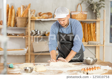Guy works in bakery as baker, kneads dough, works with flour. 125810758