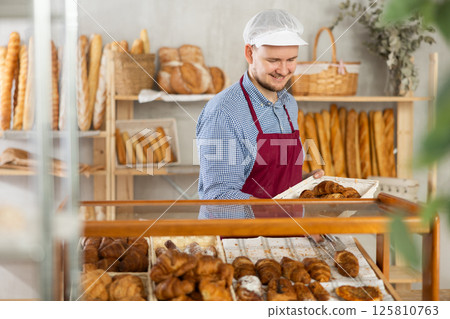 Man employee puts croissants in window, arranges display of goods at bakery. 125810763