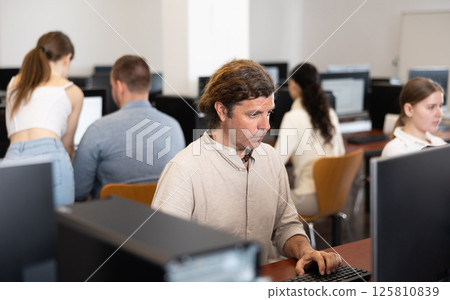 Man student of computer courses looks at monitor screen, types on keyboard and does work 125810839