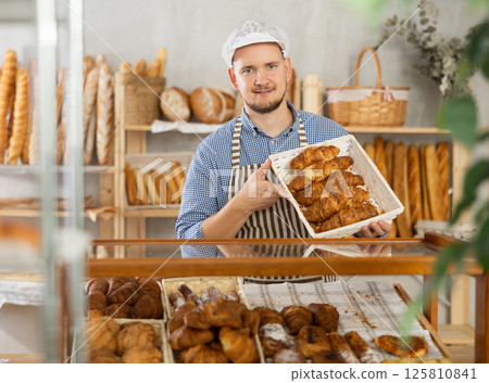 Man seller stands at bakery counter with basket filled with fresh croissants. Man seller stands at bakery counter with basket filled with fresh croissants. 125810841
