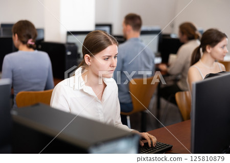 Young woman student of computer courses looks at monitor screen, types on keyboard and does work Young woman student of computer courses looks at monitor screen, types on keyboard and does work 125810879