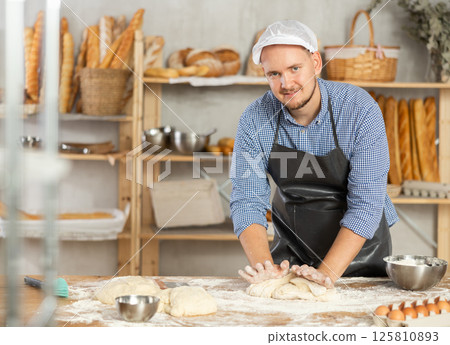 Professional baker prepares raw yeast dough in a bakery, shaping it into baguette 125810893