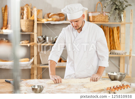 Guy works as baker in bakery, molds sausages from dough, rolls long round strips from dough 125810938