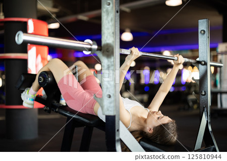 Girl lies on bench and lifts barbell, strengthens muscles of upper body Girl lies on bench and lifts barbell, strengthens muscles of upper body 125810944