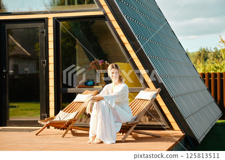 Barefoot woman sitting on folding wooden sun lounger on terrace of triangular cabin 125813511