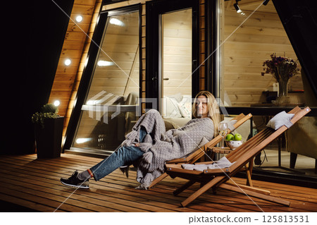 Woman wrapped in plaid enjoys fresh evening air on terrace of log cabin 125813531