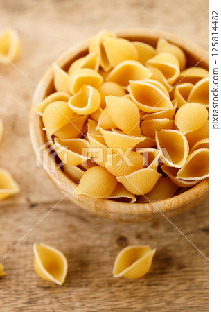 Raw uncooked dried Conchiglie shell pasta. Italian seashells pasta in wooden bowl on wooden background. 125814482