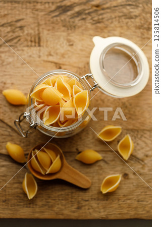 Raw uncooked dried Conchiglie shell pasta. Italian seashells pasta in lock lid glass jar on wooden background. Raw uncooked dried Conchiglie shell pasta. Italian seashells pasta in lock lid glass jar on wooden background. 125814506