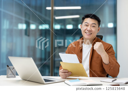 An asian man is happy and celebrating a success, holding documents at his desk. A laptop and pen holder are visible. The background shows a modern office. An asian man is happy and celebrating a success, holding documents at his desk. A laptop and pen holder are visible. The background shows a modern office. 125815648
