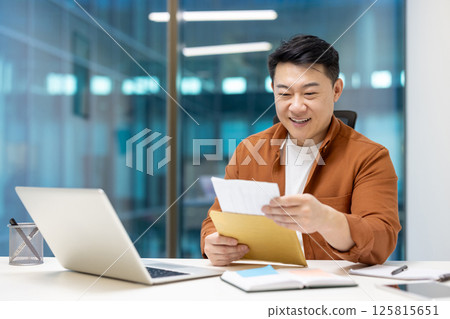 Happy Asian businessman receiving exciting mail at office, grinning while reading message. Professional workplace setting, laptop and documents present. Expression of success and enjoyment at work. 125815651