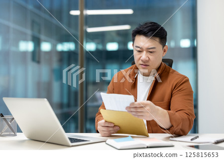 Asian man dressed casually examining documents at a modern office desk, with a laptop, notebook, and stationary, portraying a professional and focused demeanor. Asian man dressed casually examining documents at a modern office desk, with a laptop, notebook, and stationary, portraying a professional and focused demeanor. 125815653