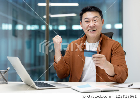 An excited asian man holds a credit card, celebrating a successful online transaction at his desk with a laptop. 125815661