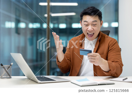 Asian man showing excitement while using a credit card for an online transaction in a contemporary office setting, showcasing joy and financial achievement. 125815666