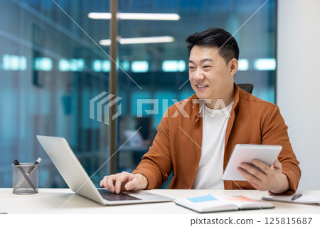 Asian businessman working on laptop and holding tablet in modern office signifying productivity, multitasking, and technology use. Reflects concepts of business, communication, and success. 125815687