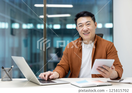 Asian man mature businessman working in an office using a tablet and laptop, smiling confidently. The atmosphere reflects productivity, success, and a positive attitude. Asian man mature businessman working in an office using a tablet and laptop, smiling confidently. The atmosphere reflects productivity, success, and a positive attitude. 125815702