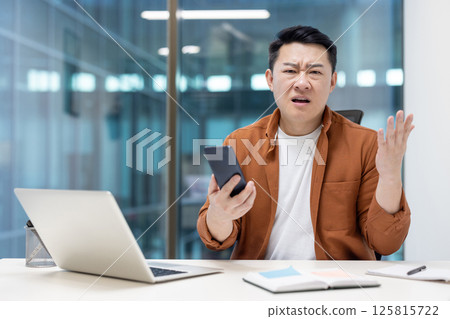 Asian man displaying frustration in a modern office, seated at a desk with a smartphone in hand, showing emotions of concern and confusion, surrounded by work materials and professional tools. 125815722