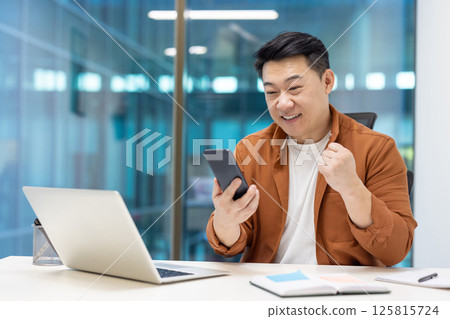 Asian businessman showing excitement looking at phone at office desk. Celebrating success in professional environment with modern tech tools around. Positive energy and motivation. Asian businessman showing excitement looking at phone at office desk. Celebrating success in professional environment with modern tech tools around. Positive energy and motivation. 125815724