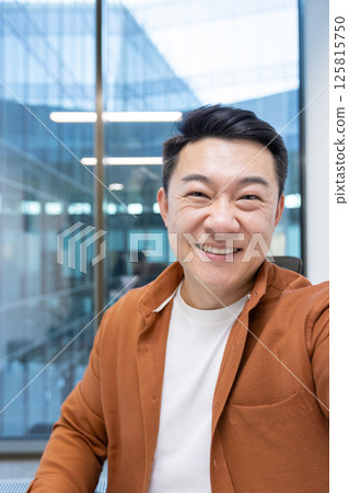 An Asian mature businessman takes a selfie video call with his phone, smiling confidently in a modern office environment, expressing professionalism and a positive demeanor. 125815750