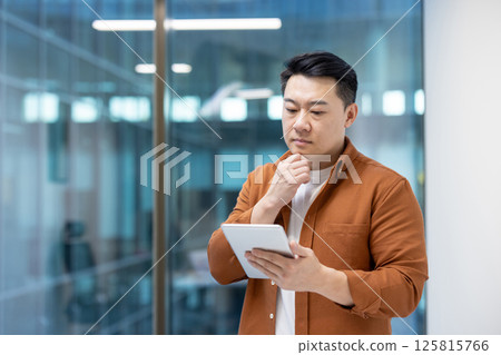 Serious thinking Asian man inside office using tablet computer. Businessman in casual clothes standing near window. 125815766