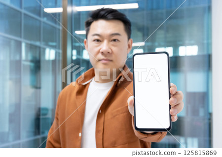 An Asian businessman holding out a smartphone with a blank screen, ideal for showcasing apps or promotions. He's inside an office building. 125815869
