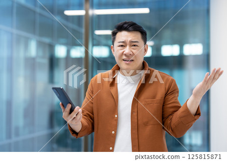 An asian man wearing an orange shirt seems to be confused by what he's reading on his mobile phone in an office setting. 125815871