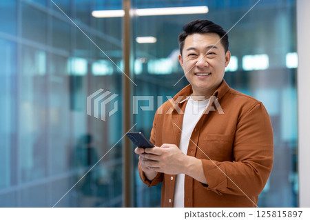 Asian businessman smiling with smartphone in hand stands confidently in modern office setting. Represents success, communication, and professional confidence in everyday business life. Asian businessman smiling with smartphone in hand stands confidently in modern office setting. Represents success, communication, and professional confidence in everyday business life. 125815897